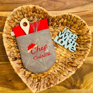 May include: A gray quilted pot holder with a red embroidered star and the text "Chef de Cuisine". The pot holder is in a woven heart-shaped basket with a wooden spoon and whisk inside. A blue wooden sign with the text "Made With Love" is also in the basket.