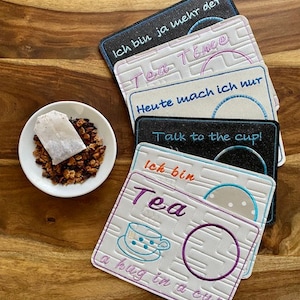 May include: Five white tea coasters with embroidered designs and text in German and English. The coasters feature a teacup, a circle, and the words "Tea", "a hug in a cup", "Talk to the cup!" and "Ich bin ja mehr der Tea Time".