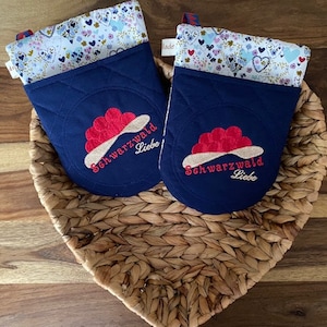 May include: Two blue quilted oven mitts with a floral pattern on the top. The mitts have red and white embroidered hearts and the text "Schwarzwald Liebe" on the front.