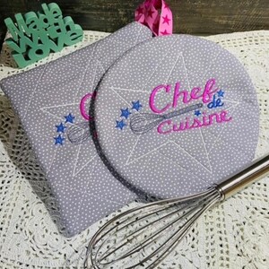 May include: Two gray quilted pot holders with white polka dots. One pot holder is round and features the text "Chef de Cuisine" embroidered in pink and blue. The other pot holder is rectangular. A silver whisk is lying on a white crocheted surface.