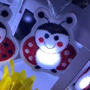 Può includere: Una ghirlanda di luci bianche con luci a forma di coccinella rossa e nera. Le luci sono infilate su un filo bianco e sono accese.