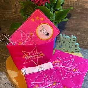 Puede incluir: Manoplas de cocina de color rosa y blanco con una estrella y el texto "Chef de Cuisine" bordado. Las manoplas están sobre una superficie de madera con una flor rosa en el fondo.
