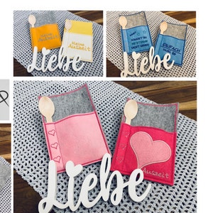 Op de afbeelding: Vier felt spoon holders met geborduurde spreuken. De houders zijn in verschillende kleuren, waaronder geel, blauw, grijs en roze. De spreuken zijn "kleine Auszeit", "SOUST KANNST DU WESTERND WICHT SEMEN", "EINFACH mal so" en "Auszeit".