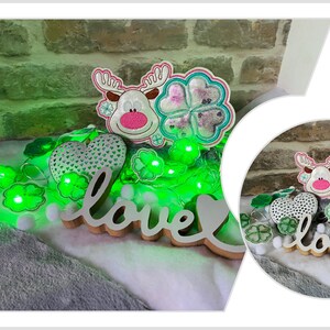 Puede incluir: Un letrero de madera blanco con la palabra "love" en cursiva, rodeado de luces verdes de trébol, un corazón blanco con acentos de trébol verde y un reno blanco con la nariz rosa y acentos de trébol verde.