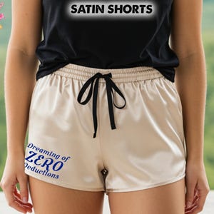 May include: Satin shorts in a light beige color with a black drawstring. The shorts have the text "SATIN SHORTS" at the top and "Dreaming of ZERO Deductions" on the left leg. The shorts are part of the "All Season's Celebrations" collection.