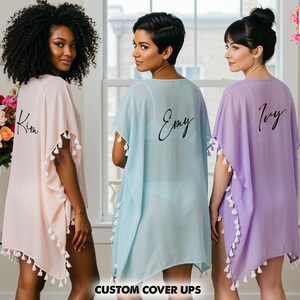 Op de afbeelding: Drie transparante cover-ups in pastelkleuren: lichtroze, lichtblauw en lavendel. Elke cover-up heeft witte kwastjes en een naam op de rug. De tekst "CUSTOM COVER UPS" staat onderaan de afbeelding. Het logo "All Season's Celebrations" staat rechtsboven.