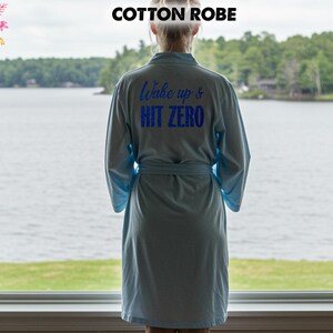 May include: A pale blue cotton robe with the text "Wake up & HIT ZERO" in blue. The robe features long sleeves and a matching belt. The image shows a person looking out a window at a lake.