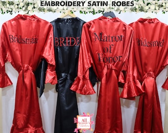 Pode incluir: Quatro batas de cetim em vermelho e preto com texto bordado. As batas têm mangas compridas com punhos com babados e um cinto para amarrar. O texto nas batas diz "Bride", "Bridesmaid", "Bridesmaid" e "Matron of Honor".
