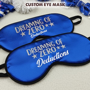 May include: Two blue satin eye masks with black elastic straps. The eye masks have silver glitter text that reads "Dreaming of Zero** Deductions".