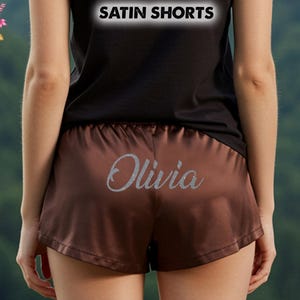 May include: Brown satin shorts with the name "Olivia" in silver glitter script. The shorts have a comfortable, relaxed fit. The top of the image reads "SATIN SHORTS". The shorts are part of the "All Season's Celebrations" collection.