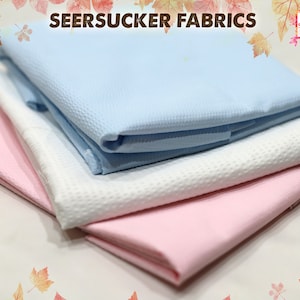 May include: Three folded pieces of seersucker fabric in different colors: light blue, white, and pink. The fabric has a textured, crinkled surface.