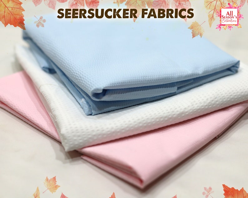 May include: Three folded pieces of seersucker fabric in different colors: light blue, white, and pink. The fabric has a textured, crinkled surface.
