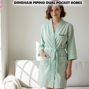 Op de afbeelding: Lichtgroene geruite badjas met witte bies, twee zakken en een bijpassende riem. De badjas heeft de naam "Sandra" op de borst geborduurd. De tekst "GINGHAM PIPING DUAL POCKET ROBES" staat bovenaan de afbeelding.