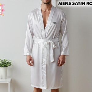 Può includere: Un accappatoio di raso bianco con cintura annodata e maniche lunghe. La parola "Max" è ricamata sul petto. Il testo "MENS SATIN ROBES" è visualizzato nella parte superiore dell'immagine. Sono visibili anche una piccola pianta e una sveglia.