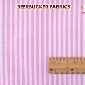 May include: Pink and white striped seersucker fabric. The fabric has vertical stripes and a textured appearance. A wooden ruler is placed on the fabric, showing measurements in centimeters. The text "SEERSUCKER FABRICS" is at the top.