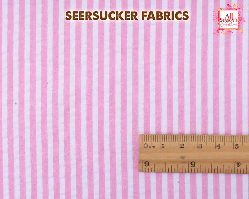 May include: Pink and white striped seersucker fabric. The fabric has vertical stripes and a textured appearance. A wooden ruler is placed on the fabric, showing measurements in centimeters. The text "SEERSUCKER FABRICS" is at the top.