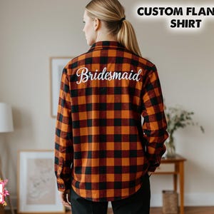 May include: A custom flannel shirt in a classic orange and black plaid pattern. The word "Bridesmaid" is embroidered in white script across the back. The top right corner of the image has the text "CUSTOM FLANNEL SHIRT".