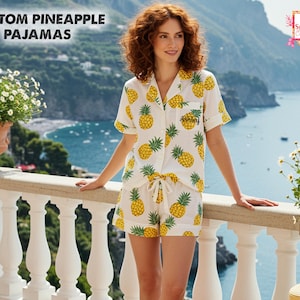 May include: White pajama set with a yellow pineapple pattern. The set includes a short-sleeved button-down shirt and shorts. The text "Custom Pineapple Pajamas" is at the top of the image. The shirt pocket has the name "Ashley" embroidered on it.