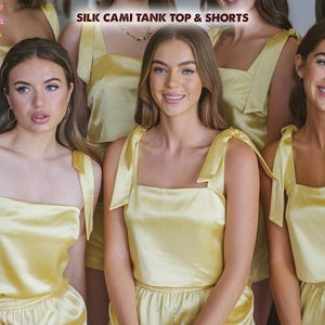 May include: Four women wearing matching yellow silk cami tank tops and shorts. The tops have thin straps with a bow detail. The shorts have an elastic waistband. The text "SILK CAMI TANK TOP & SHORTS" is displayed at the top of the image.