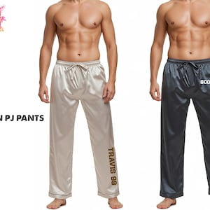 May include: Satin pajama pants in cream and charcoal gray. The cream-colored pants have "TRAVIS 99" printed on the leg. The charcoal gray pants have "SCOTT" printed on the leg. Both pairs have a drawstring waist and are labeled "SATIN PJ PANTS".