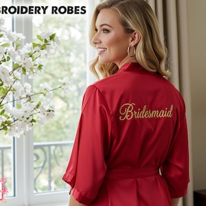 Op de afbeelding: Een rode satijnen badjas met het woord "Bridesmaid" in gouden letters geborduurd. De badjas heeft een bijpassende riem en maakt deel uit van een collectie "Embroidery Robes". De afbeelding toont ook een raam met witte bloemen en het logo "All Season's Celebrations".