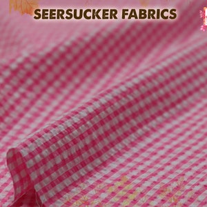 May include: Pink and white seersucker fabric with a gingham pattern. The fabric is folded and has a slight crinkle texture.
