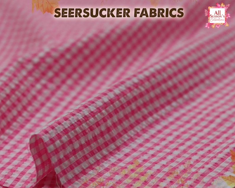 May include: Pink and white seersucker fabric with a gingham pattern. The fabric is folded and has a slight crinkle texture.