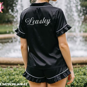 May include: Black satin pajama set with white trim and ruffle detailing. The short-sleeved top and shorts feature the name "Leasley" in white script on the back. The words "Ruffle Satin Pajamas" are at the bottom of the image.