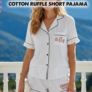 May include: White cotton pajama set with black trim, featuring a short-sleeved top with ruffles and a pocket with the name "Holly". The matching shorts have the text "Cheer Squad". The top has black buttons.