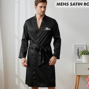 May include: A black satin robe with a shawl collar and a matching tie belt. The robe features long sleeves and the name "Max" embroidered on the chest. The text "MENS SATIN ROBES" is at the top of the image.