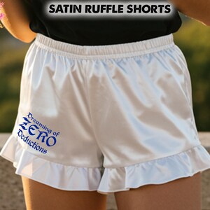 May include: White satin ruffle shorts with an elastic waistband and a frilled hem. The shorts feature the text "Dreaming of ZERO Deductions" in blue. The words "SATIN RUFFLE SHORTS" are at the top of the image.