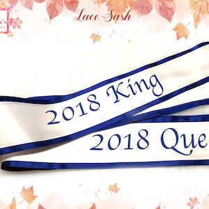 May include: White satin sash with blue trim and the text "2018 King" and "2018 Queen" in blue lettering. The sash is a lace sash.