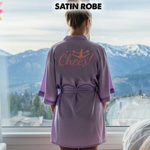 May include: A lilac satin robe with the word "Cheer!" and a cheerleader graphic printed on the back. The robe has a matching belt and trim. The text "SATIN ROBE" is at the top of the image.