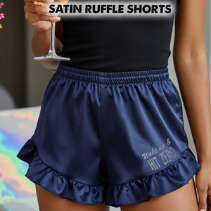 May include: Navy blue satin ruffle shorts with a frilled hem. The shorts have the text "Wake up & HIT ZERO" embroidered on the front. The image also includes the text "SATIN RUFFLE SHORTS".