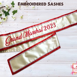 May include: A gold satin sash with red trim and rhinestones. The sash is embroidered with the text "Grand Marshal 2023".