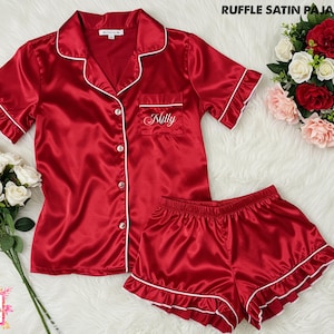 May include: Red satin pajama set with white trim, featuring a button-down shirt and shorts. The shirt has a pocket with the name "Milly" embroidered. The set includes ruffle detailing on the sleeves and shorts. The text "Ruffle Satin Pajamas" is visible.