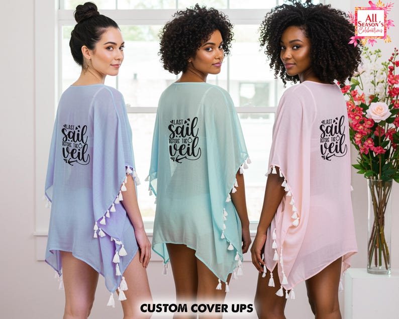 Op de afbeelding: Drie vrouwen dragen doorschijnende cover-ups in pasteltinten: lavendel, mintgroen en roze. Elke cover-up heeft witte kwastjes en de tekst "Last Sail Before The Veil" op de rug. De cover-ups zijn licht en luchtig.