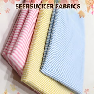May include: Three different colors of seersucker fabric, pink, yellow, and blue, are stacked on top of each other. The fabric is striped and has a crinkled texture.