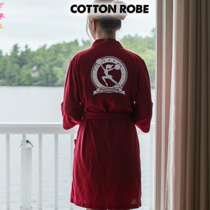 May include: A burgundy cotton robe with a white "Cheerleading" logo on the back. The robe is tied at the waist and has long sleeves. The text "COTTON ROBE" is at the top of the image. The robe is worn by a person standing near a window.