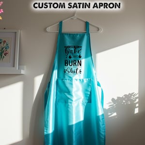 May include: A custom satin apron in a vibrant turquoise color, hanging on a white hanger. The apron features a large pocket and the text "I like to bake and burn shit" in black. The apron is displayed against a white wall.