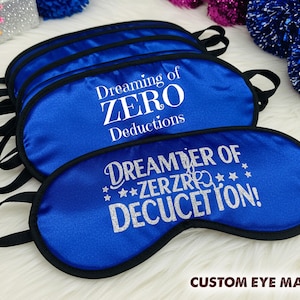 May include: A collection of blue satin eye masks with black trim and elastic straps. The masks feature white text: "Dreaming of ZERO Deductions" and "DREAMBER OF ZERZRRO DECUCETTON!". The words "CUSTOM EYE MASK" are also visible.