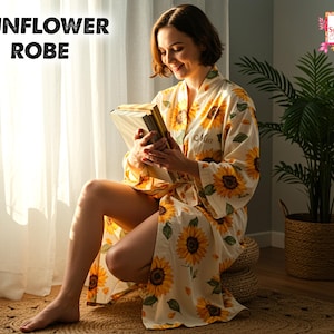 Könnte beinhalten: Ein cremefarbener Sonnenblumen-Bademantel mit gelbem Sonnenblumenmuster. Der Bademantel hat lange Ärmel und einen Bindverschluss. Der Text "Sunflower Robe" steht oben links. Der Bademantel ist mit dem Namen "Mia" personalisiert.