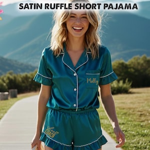 Puede incluir: Conjunto de pijama corto de satén verde azulado con ribete de volantes. La parte superior de manga corta y los pantalones cortos tienen ribetes blancos y un nombre personalizado bordado en el bolsillo. El texto "Satin Ruffle Short Pajama" está en la parte superior de la imagen.