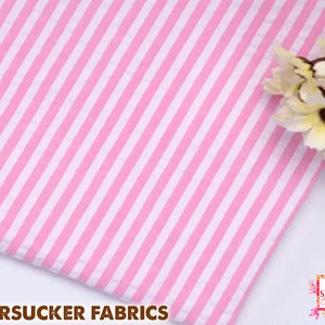 May include: Pink and white striped seersucker fabric. The fabric has vertical stripes of alternating pink and white. The words "SEERSUCKER FABRICS" are printed at the bottom of the image.