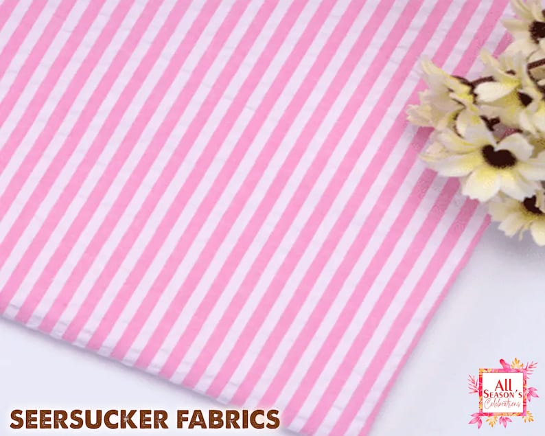 May include: Pink and white striped seersucker fabric. The fabric has vertical stripes of alternating pink and white. The words "SEERSUCKER FABRICS" are printed at the bottom of the image.