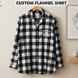 Può includere: Una camicia di flanella a quadri bianchi e neri con il nome "Albert" ricamato sulla tasca. La camicia abbottonata a maniche lunghe è appesa a una gruccia di legno. Il testo "CUSTOM FLANNEL SHIRT" è sopra.