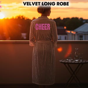 May include: A light-coloured velvet long robe with the word "CHEER" in pink lettering on the back. The robe has a matching belt and is worn by a person standing on a balcony at sunset. A small table with glasses and a carafe is nearby.