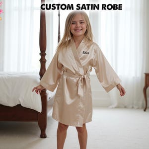 May include: A champagne-colored satin robe featuring wide sleeves, a matching tie belt at the waist, and the name Eden embroidered in black script on the left chest. The text CUSTOM SATIN ROBE is displayed at the top of the image.