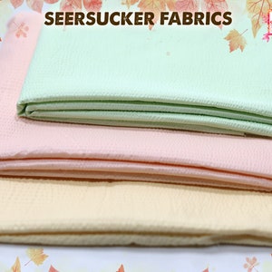 May include: Three different colors of seersucker fabric, folded and stacked on top of each other. The colors are light green, pink, and light yellow.