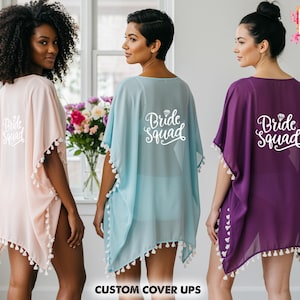 Op de afbeelding: Drie transparante cover-ups in roze, blauw en paars, elk met witte kwastjes en de woorden "Bride Squad" op de rug gedrukt. De cover-ups worden getoond op drie vrouwen voor een raam met bloemen.